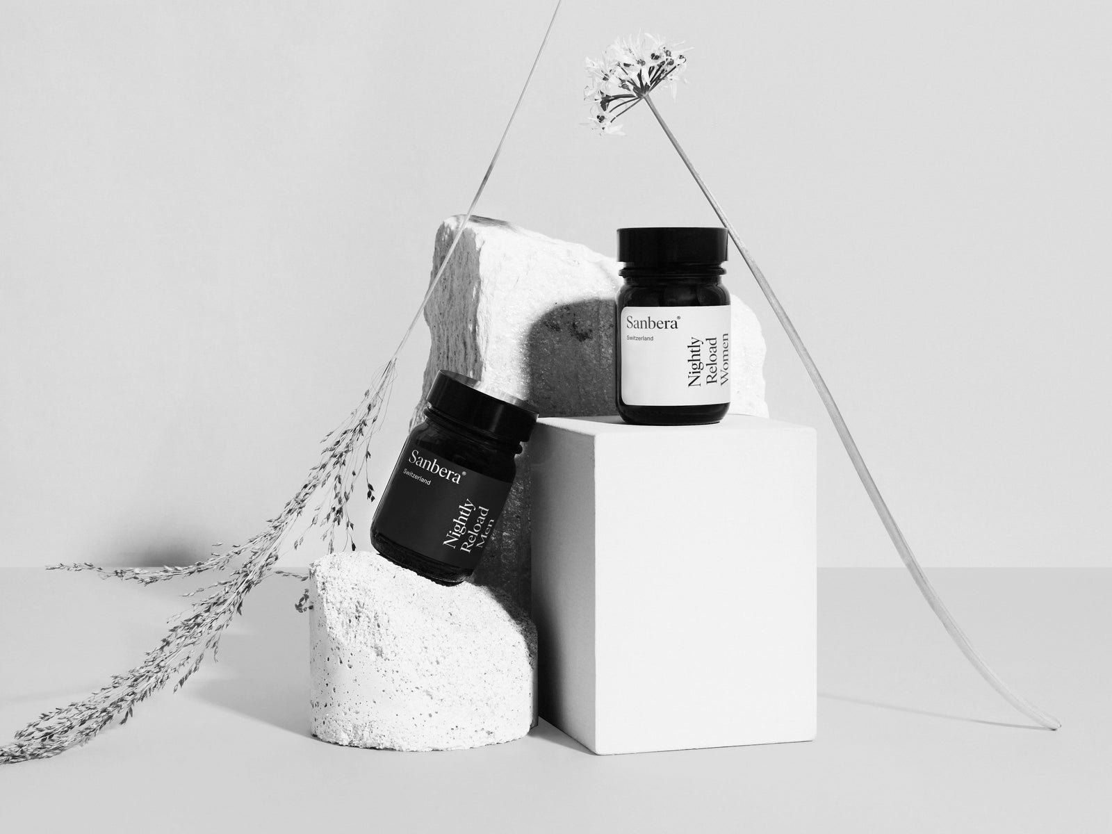Sanbera Nightly Reload Women and Men supplement bottles displayed on stone blocks with delicate botanical stems.